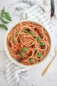 Vegan spaghetti with spicy tomato sauce, black olives, and fresh basil leaves served in a white bowl on a marble surface