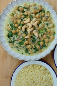 Creamy coconut chickpea curry with kale and cashew topping, served with a bowl of fluffy couscous on a wooden surface.