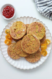 Baked zucchini and tofu burgers served on a white plate with yellow cherry tomatoes and a side of ketchup